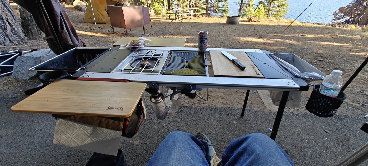 A camp kitchen setup with a built-in stove, griddle, and cutting boards at a lakeside campsite