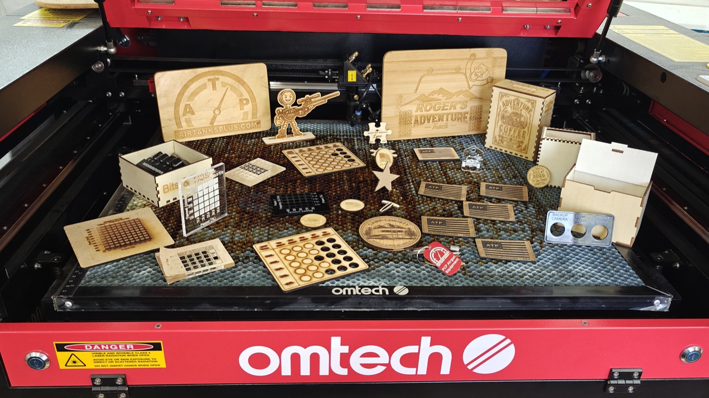 A laser cutter bed full of finished wood projects — AirTanksPlus signs, Adventure Truck panels, tags, and small boxes