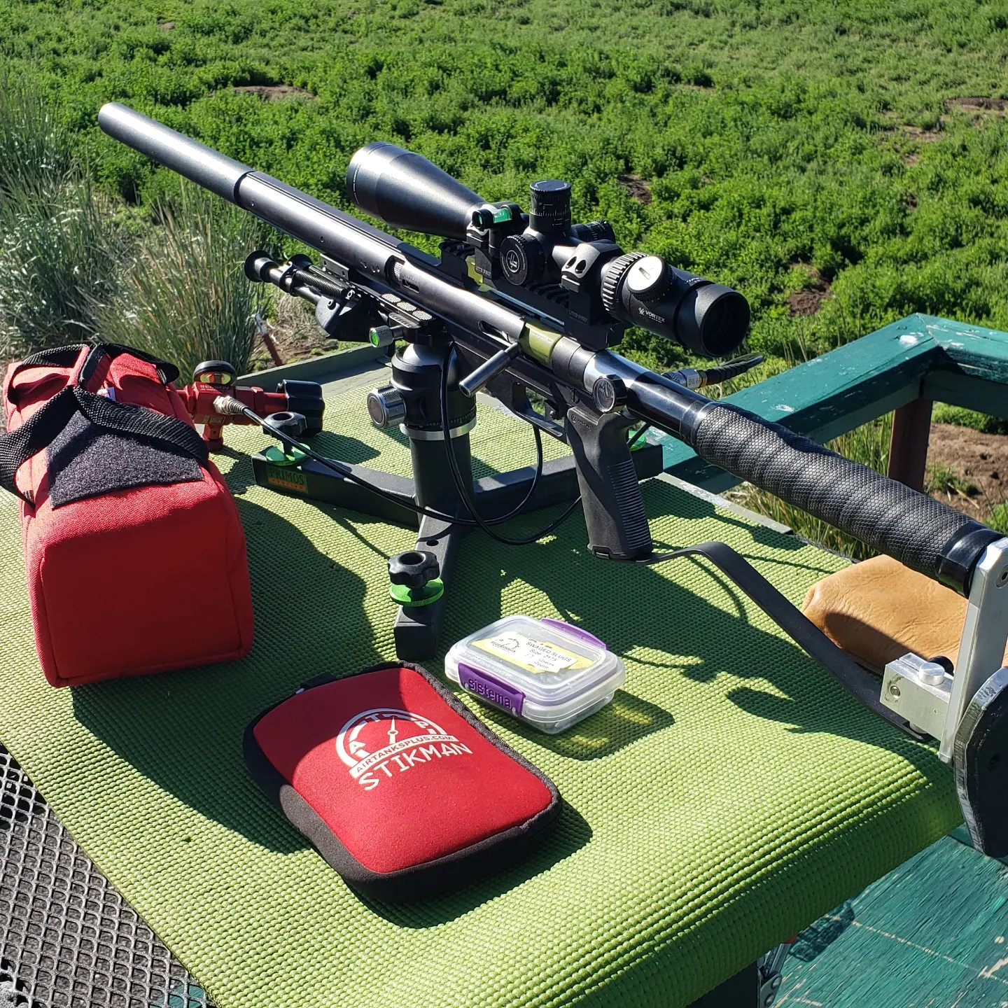 A scoped PCP air rifle on a shooting rest at the bench, with a STIKMAN fill system pouch and ammo case nearby
